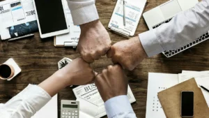 Mãos de quatro pessoas unidas sobre mesa de escritório com documentos e notebooks, representando a parceria entre contratante e prestadora de serviços