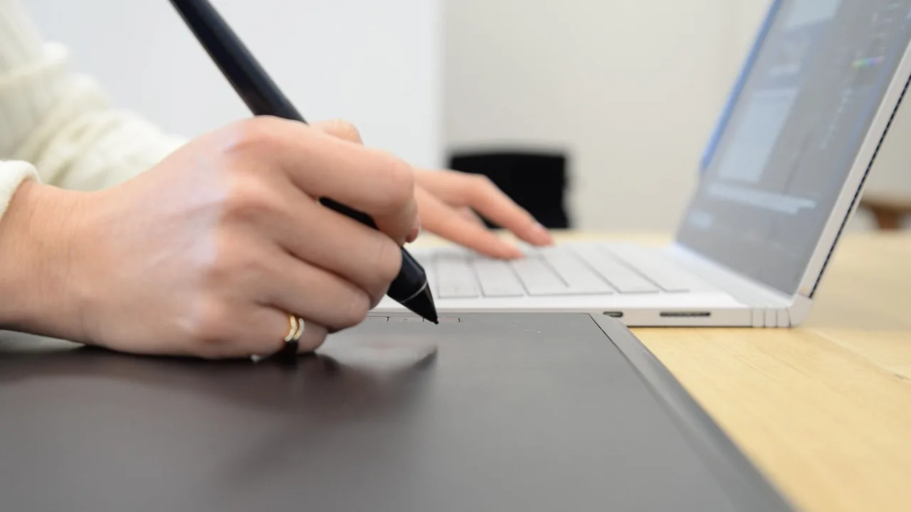 Mão usando caneta em mesa digitalizadora ao lado de notebook, representando a criação de peças por uma agência parceira