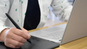 Mãos desenhando com caneta em mesa digitalizadora ao lado de notebook, representando o trabalho de um ilustrador gráfico