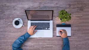 Mãos em notebook e mesa digitalizadora vistas de cima, representando o trabalho de criação da agência