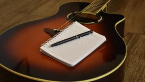 Violão sobre piso de madeira com caderno e caneta, representando a composição de uma música