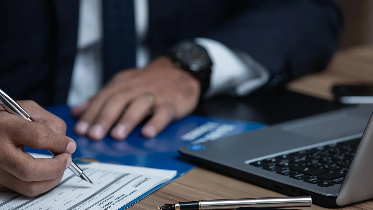 Mãos assinando documento ao lado de notebook, representando a assinatura de contrato entre empresas