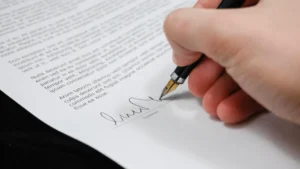 Mão assinando contrato com caneta-tinteiro sobre a mesa, representando o contrato de prestação de serviços
