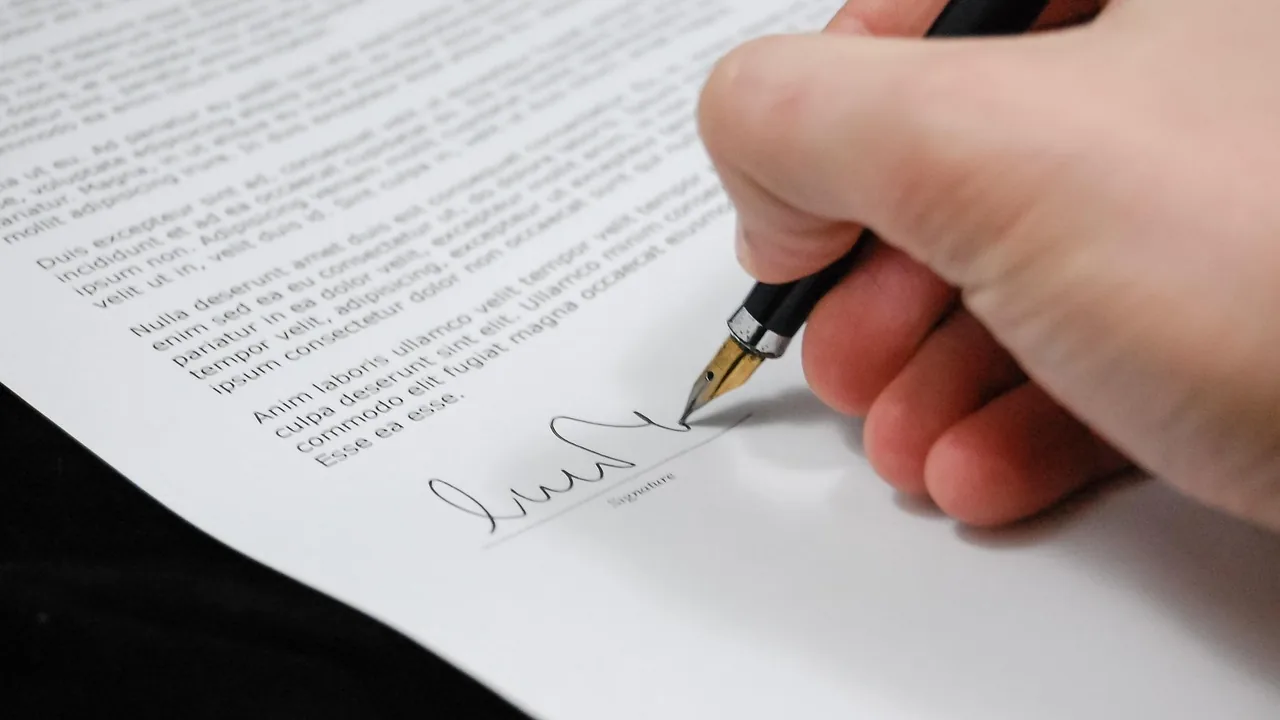 Mão assinando contrato com caneta-tinteiro sobre a mesa, representando o contrato de prestação de serviços