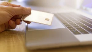 Mão segurando cartão sem marca ao lado de um notebook, representando o pagamento em loja virtual